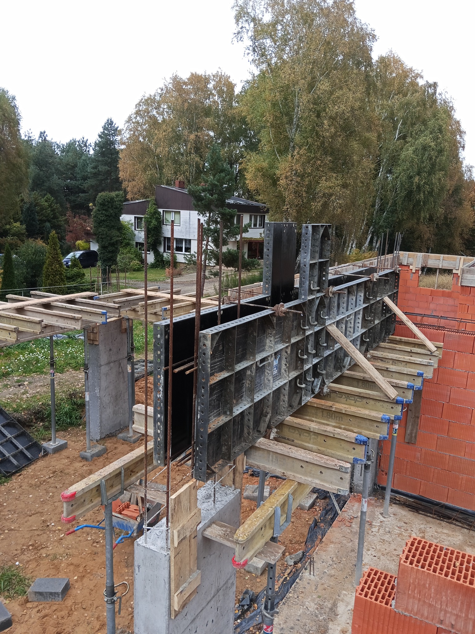 Wysoka perspektywa na szalunek fundamentów z metalowymi panelami i drewnianymi belkami, częściowo zabetonowany, z widocznymi prętami zbrojeniowymi i fragmentem murowanej ściany.