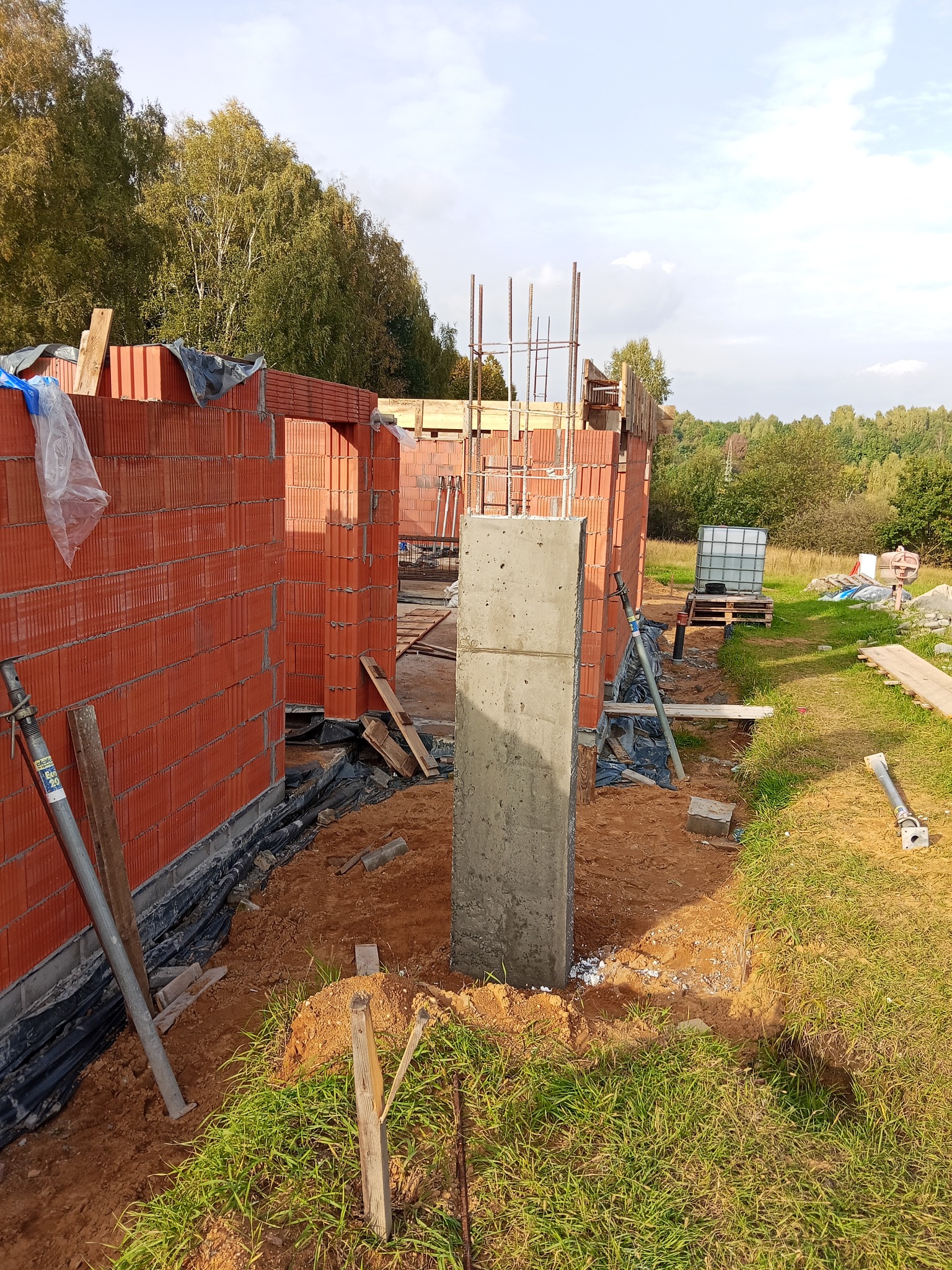 Wznoszenie ścian z czerwonej cegły na budowie domu. Widoczny betonowy filar, zbrojenie i elementy konstrukcyjne. W tle drzewa i niebo.