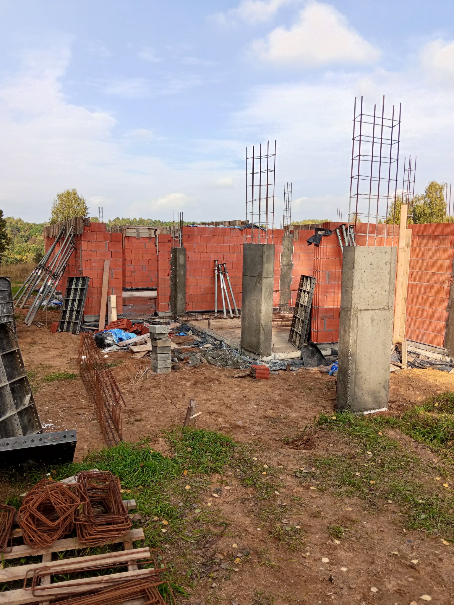 Wznoszenie ścian z czerwonej cegły i betonowych kolumn na placu budowy. Widoczne zbrojenia, szalunki i narzędzia. Etap budowy domu w otoczeniu zieleni.