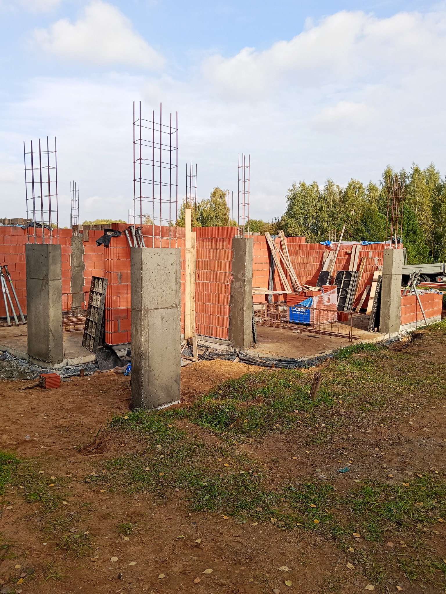 Budowa domu: ściany z czerwonej cegły, betonowe słupy i zbrojenia na tle zieleni i błękitnego nieba. Widoczne materiały budowlane i elementy konstrukcyjne.