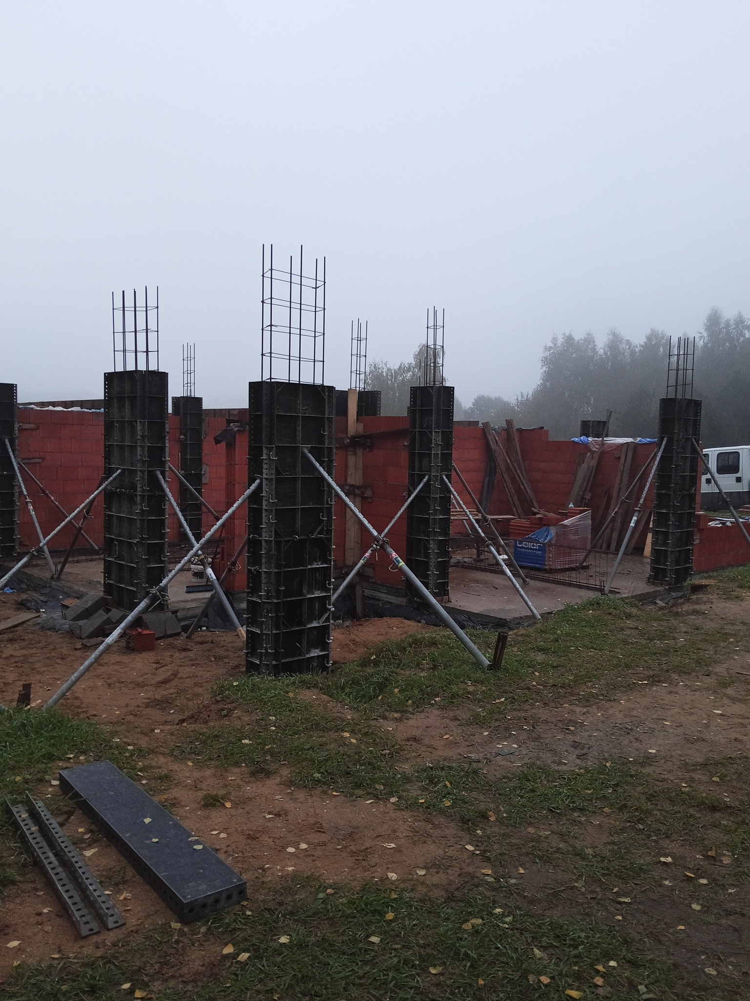 Wznoszenie konstrukcji domu: zbrojenie kolumn w szalunkach, widoczne ceglane ściany w tle, pochmurna pogoda. Elementy konstrukcyjne na placu budowy.