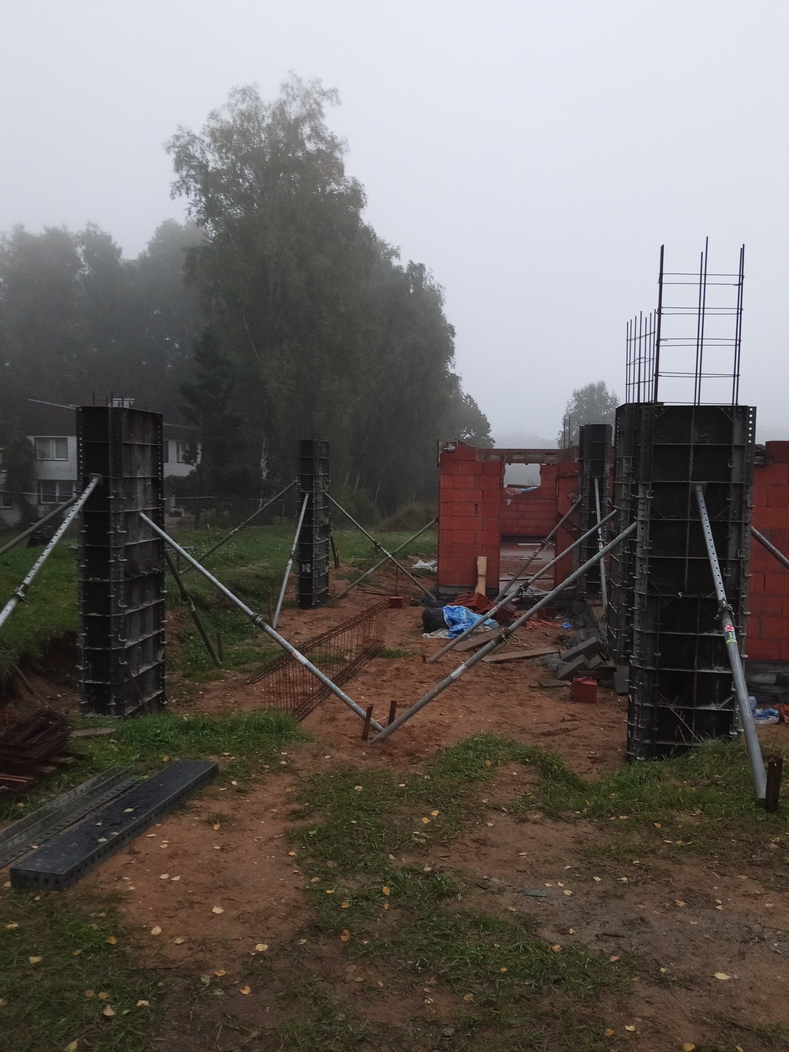 Budowa domu w trakcie: widoczne szalunki kolumnowe ze zbrojeniem, fragment muru z czerwonej cegły. Mglista pogoda, drzewa w tle. Ujęcie z poziomu gruntu.