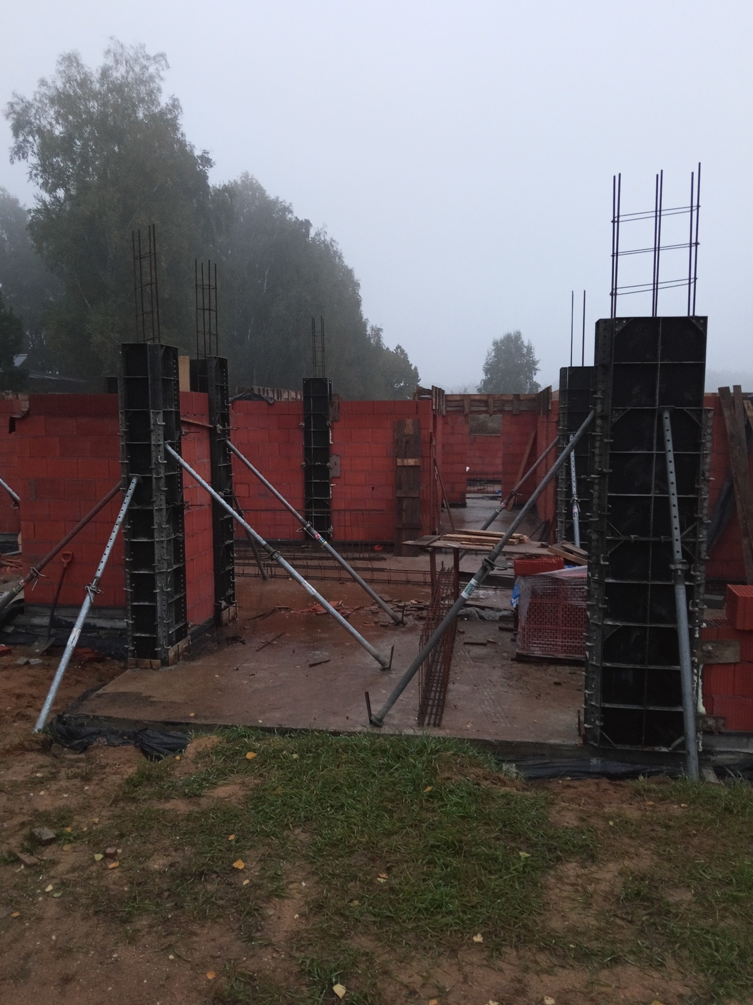 Budowa domu z czerwonej cegły: zbrojone słupy w szalunkach, wsparte metalowymi podporami. Mglista pogoda w tle. Widoczne elementy konstrukcyjne i materiały budowlane.
