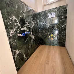 PN-BUD PAWEŁ NOWAK - Fragment łazienki w trakcie remontu: ściany wyłożone zielonym marmurem z widocznymi przyłączami wodnymi i stelażem podtynkowym WC, podłoga wyłożona panelami imitującymi drewno, surowe ściany, okno...