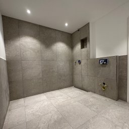 PN-BUD PAWEŁ NOWAK - Surowa łazienka w szarościach przed montażem armatury, widoczne przyłącza wodne i kanalizacyjne, wnęka w ścianie, płytki imitujące kamień na ścianach i podłodze.