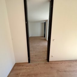 PN-BUD PAWEŁ NOWAK - Wnętrze wykończonego pokoju z panelami podłogowymi i widocznym przejściem do kolejnego pomieszczenia z ciemnymi futrynami.