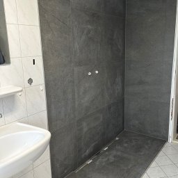 PN-BUD PAWEŁ NOWAK - Fragment łazienki po montażu szarych płytek imitujących kamień w strefie prysznicowej i na podłodze, widoczny biały zlew, białe płytki na ścianie oraz instalacja wodna bez armatury.