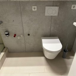 PN-BUD PAWEŁ NOWAK - Wisząca toaleta z szarą szczotką i zabudowanym systemem spłukiwania na szarej, marmurowej ścianie z gniazdkami elektrycznymi, widoczne przyłącza wodne i fragment kabiny prysznicowej.