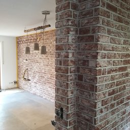 PN-BUD PAWEŁ NOWAK - Wnętrze w trakcie wykańczania z dekoracyjną ścianą z cegły, widoczne niedokończone instalacje elektryczne i surowa podłoga, z lampą wiszącą w stylu industrialnym.