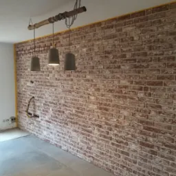 Ściana wykończona cegłą dekoracyjną w odcieniach czerwieni i bieli, z widocznymi przewodami elektrycznymi wystającymi ze ściany i trzema szarymi lampami wiszącymi na drewnianej belce, żółta taśma...