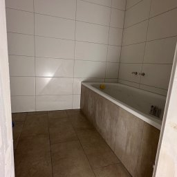 PN-BUD PAWEŁ NOWAK - Łazienka w trakcie wykończenia: ułożone brązowe płytki podłogowe i białe płytki ścienne, obudowana wanna. Widoczne niedokończone wykończenia.