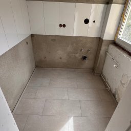 PN-BUD PAWEŁ NOWAK - Łazienka w trakcie prac: ułożona podłoga z płytek, częściowo położone białe płytki na ścianach, widoczne instalacje elektryczne i hydrauliczne. Okno z prawej strony.