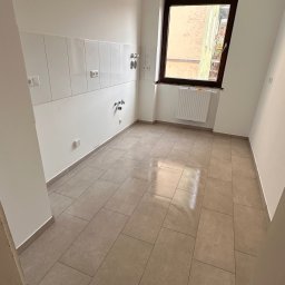 PN-BUD PAWEŁ NOWAK - Jasna łazienka po remoncie: beżowe płytki podłogowe, białe ściany z fragmentem glazury, brązowe okno. Widoczne przyłącza wodne i elektryczne.