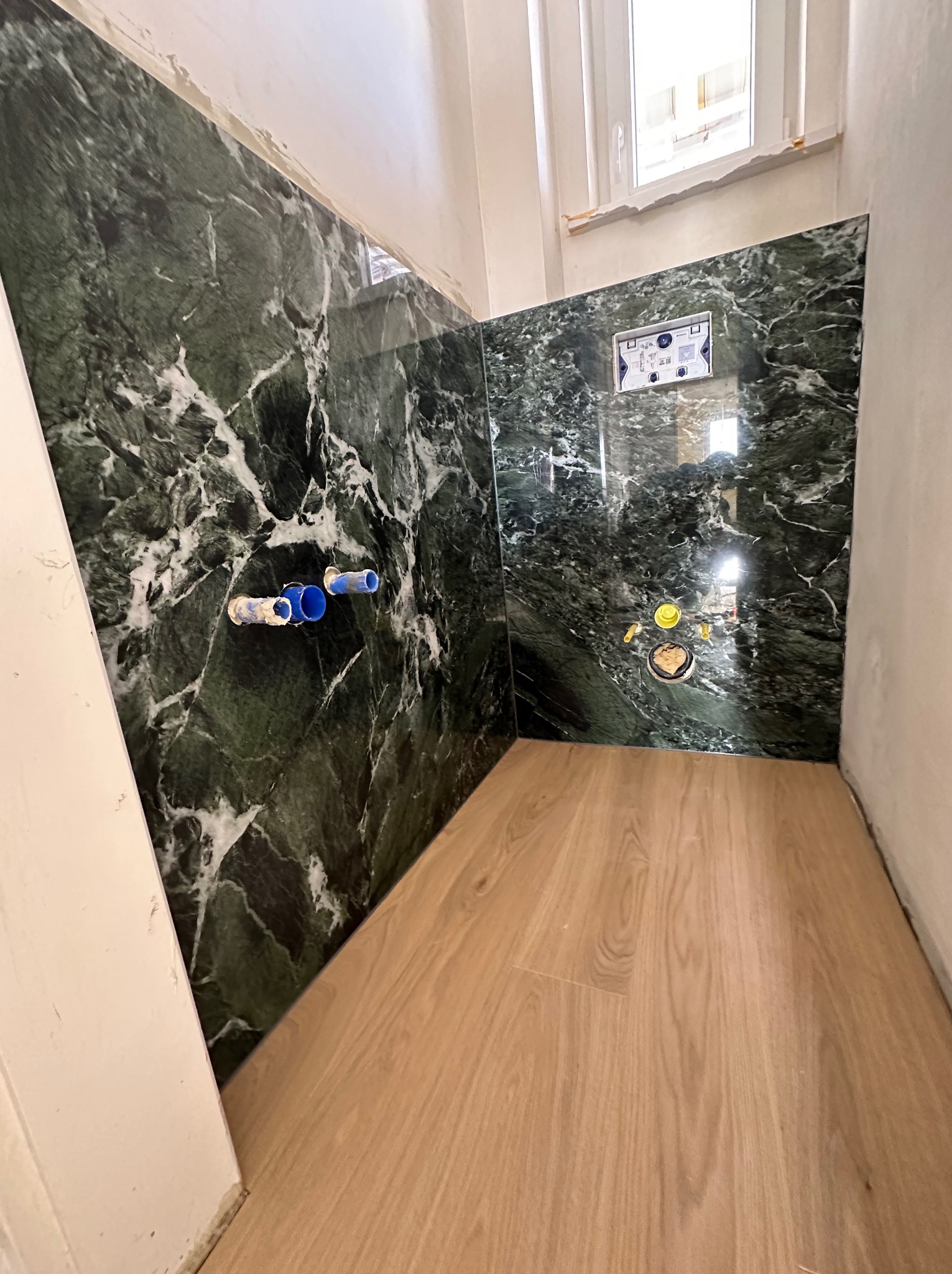 Fragment łazienki w trakcie remontu: ściany wyłożone zielonym marmurem z widocznymi przyłączami wodnymi i stelażem podtynkowym WC, podłoga wyłożona panelami imitującymi drewno, surowe ściany, okno...