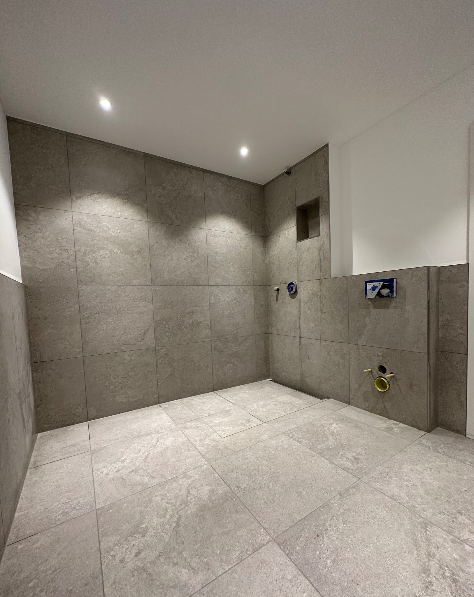 Surowa łazienka w szarościach przed montażem armatury, widoczne przyłącza wodne i kanalizacyjne, wnęka w ścianie, płytki imitujące kamień na ścianach i podłodze.