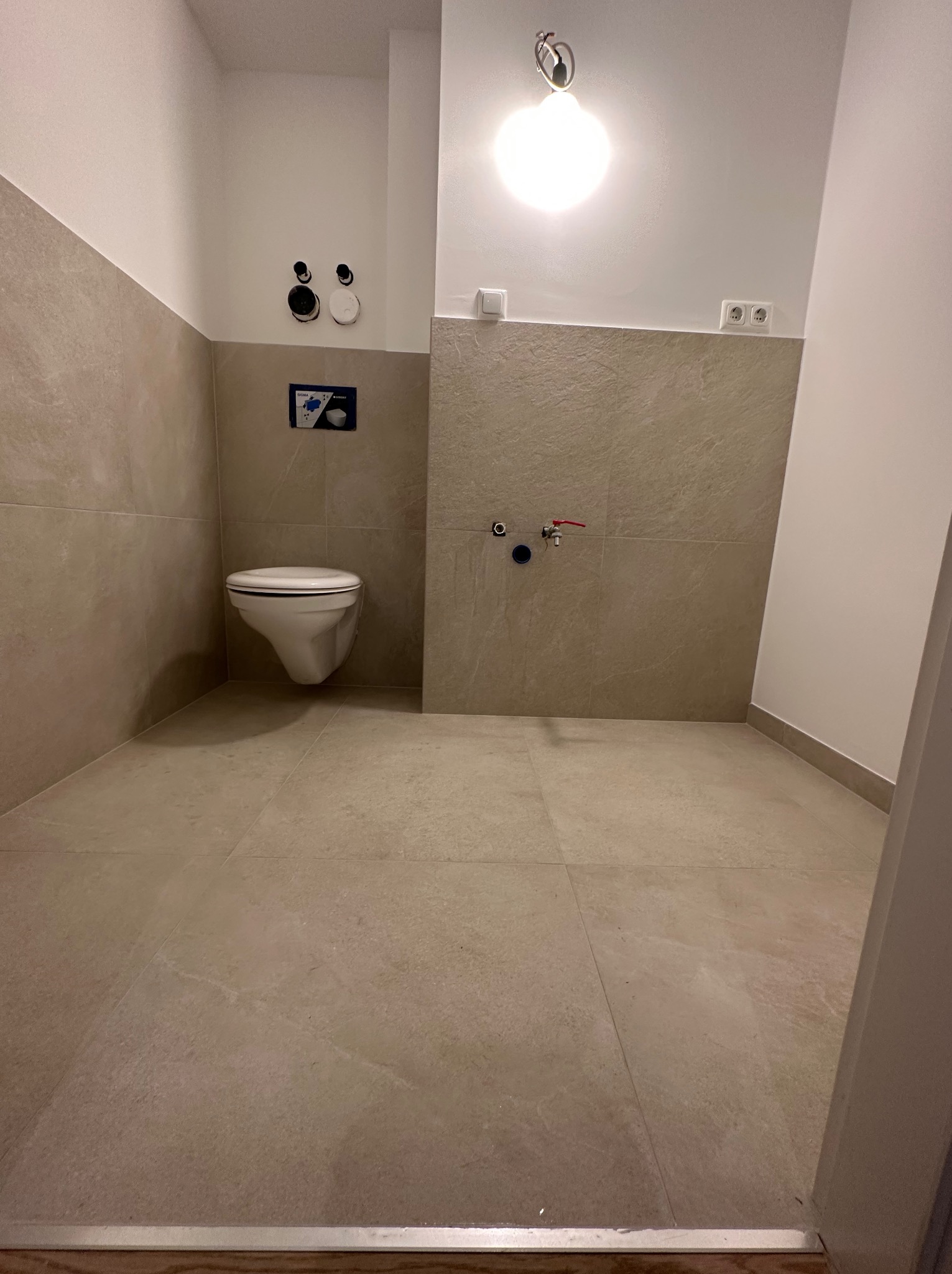 Nowoczesna łazienka z podwieszaną toaletą, wykończona beżowymi płytkami, widoczne przyłącza wodne i kanalizacyjne, minimalistyczne oświetlenie sufitowe.