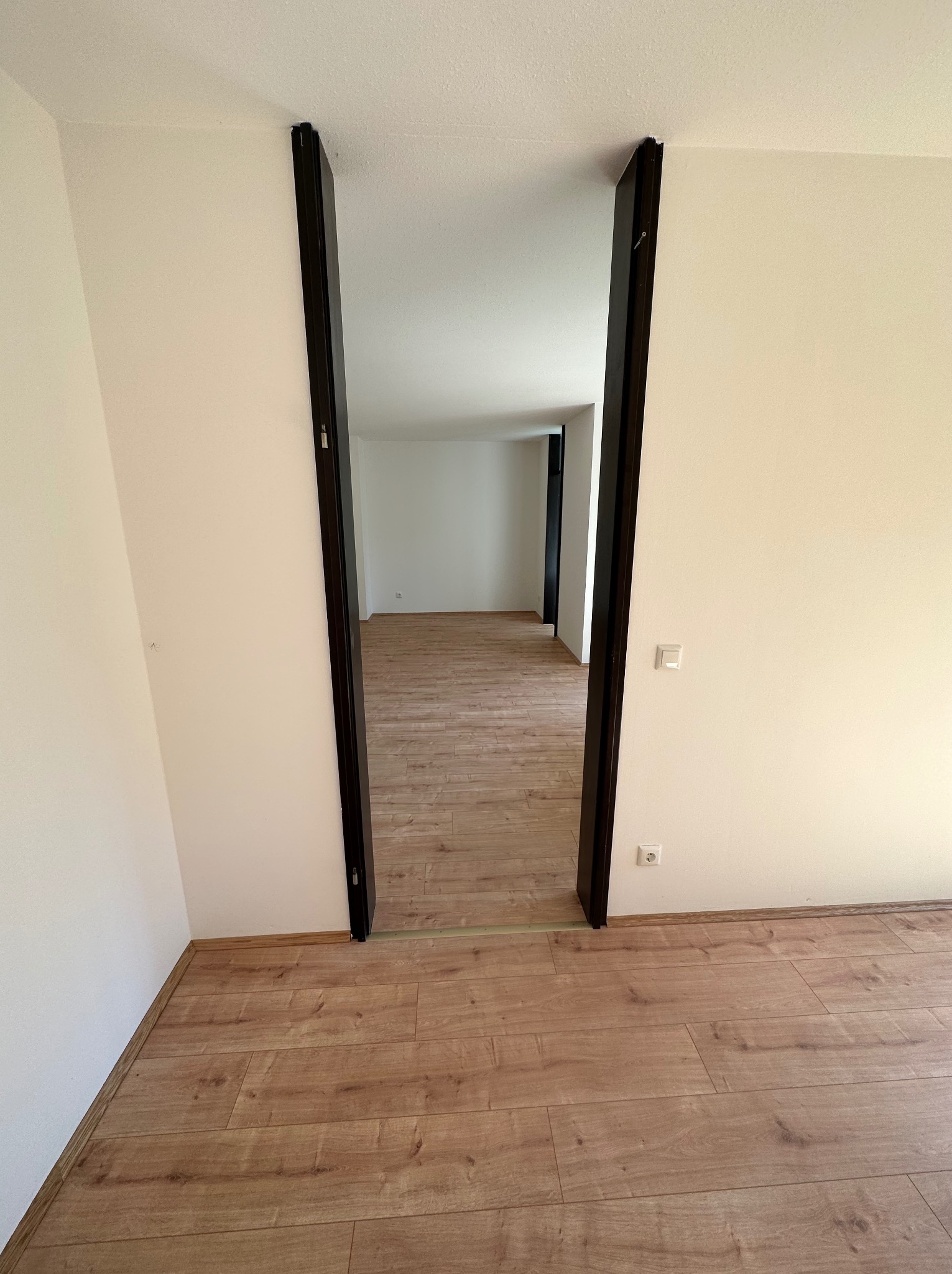 Wnętrze wykończonego pokoju z panelami podłogowymi i widocznym przejściem do kolejnego pomieszczenia z ciemnymi futrynami.