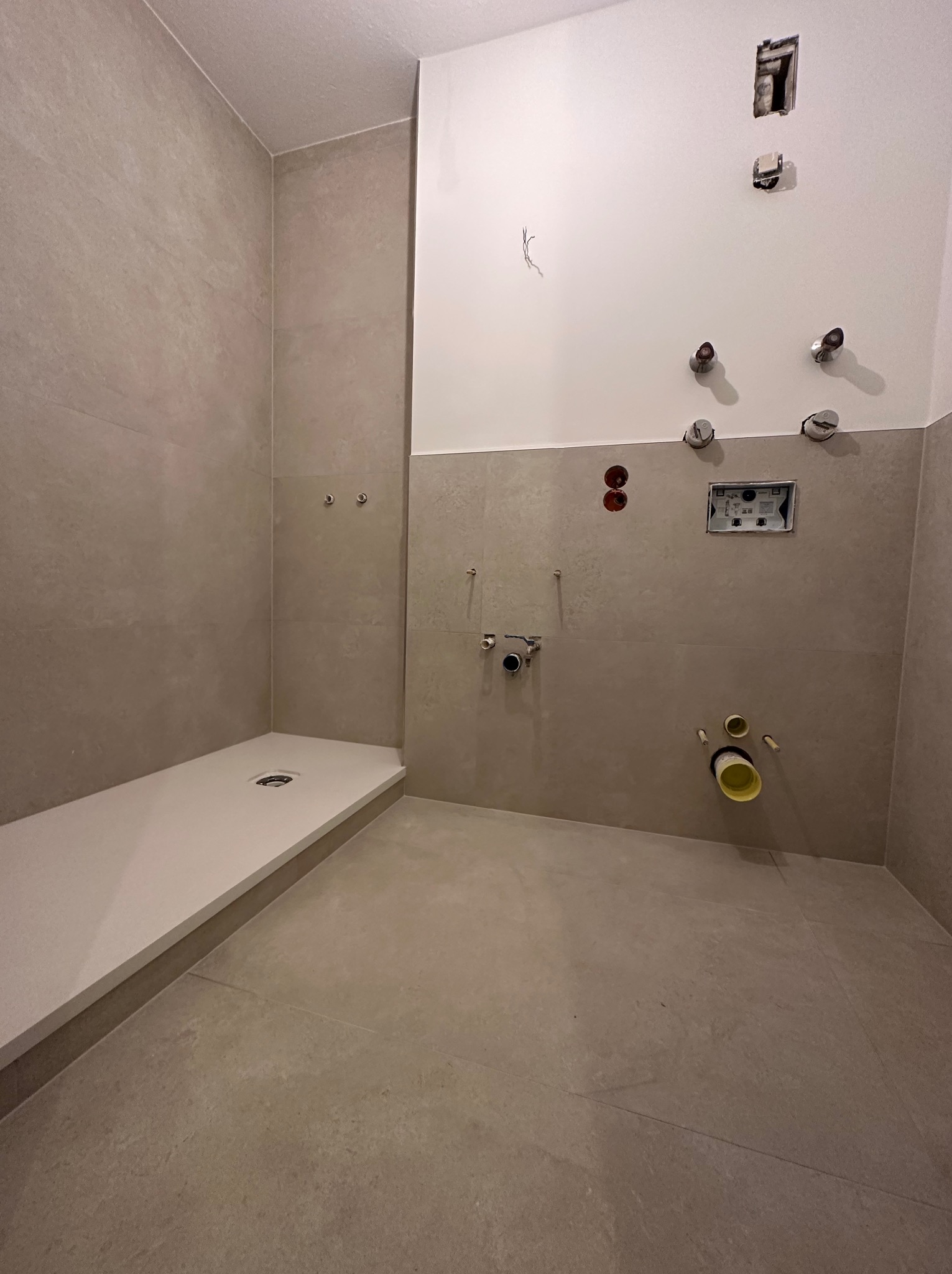 Wnętrze łazienki w trakcie remontu: szara glazura na ścianach i podłodze, biały brodzik, wystające rury i instalacje wodno-kanalizacyjne, otwór w ścianie po instalacji elektrycznej.