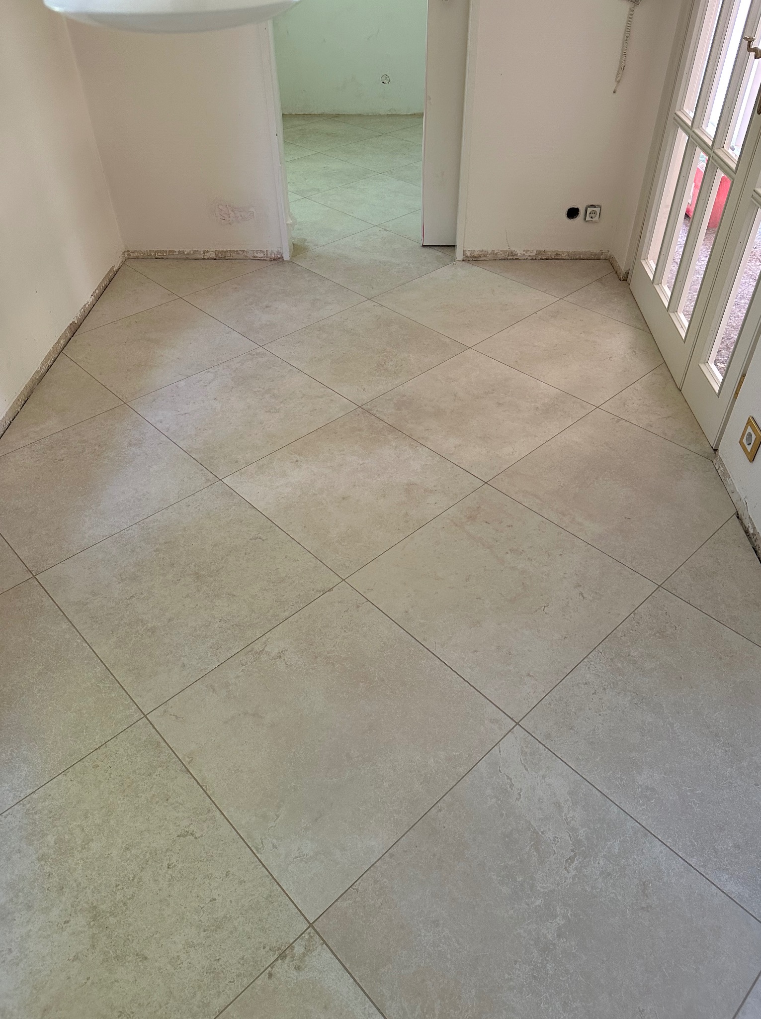 Beżowe płytki podłogowe ułożone diagonalnie w jasnym pomieszczeniu, widoczne ślady po usuniętych listwach przypodłogowych i gniazdka elektryczne na ścianie.