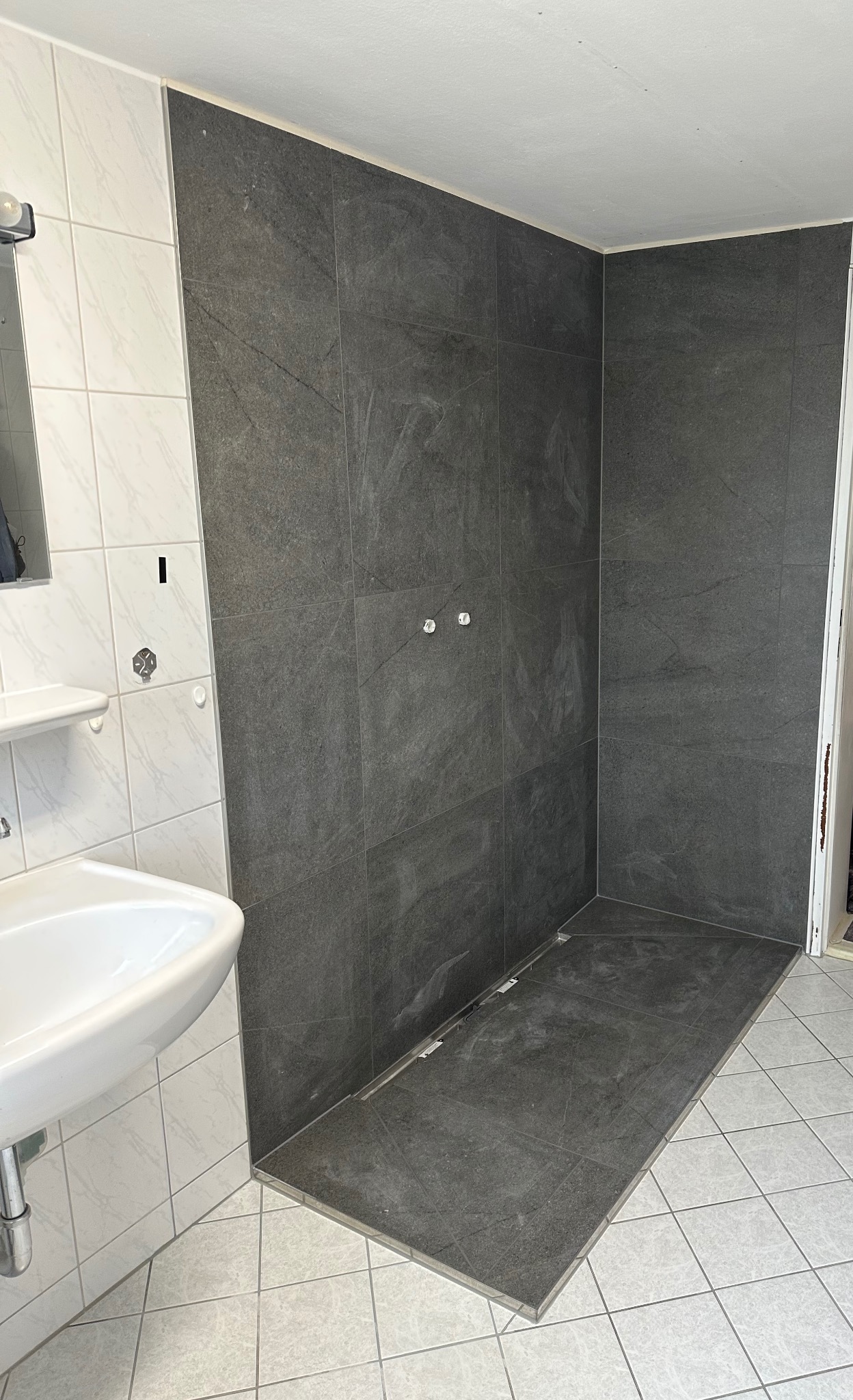 Fragment łazienki po montażu szarych płytek imitujących kamień w strefie prysznicowej i na podłodze, widoczny biały zlew, białe płytki na ścianie oraz instalacja wodna bez armatury.