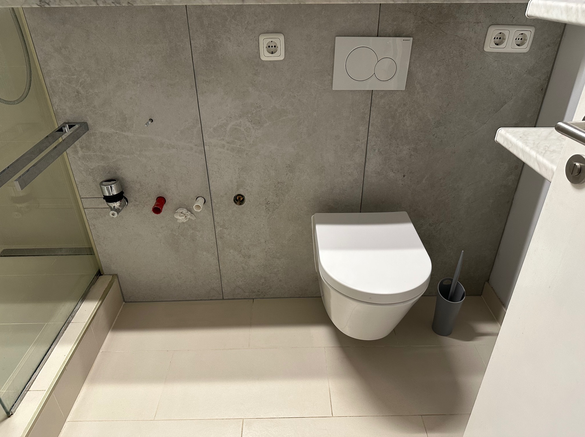 Wisząca toaleta z szarą szczotką i zabudowanym systemem spłukiwania na szarej, marmurowej ścianie z gniazdkami elektrycznymi, widoczne przyłącza wodne i fragment kabiny prysznicowej.