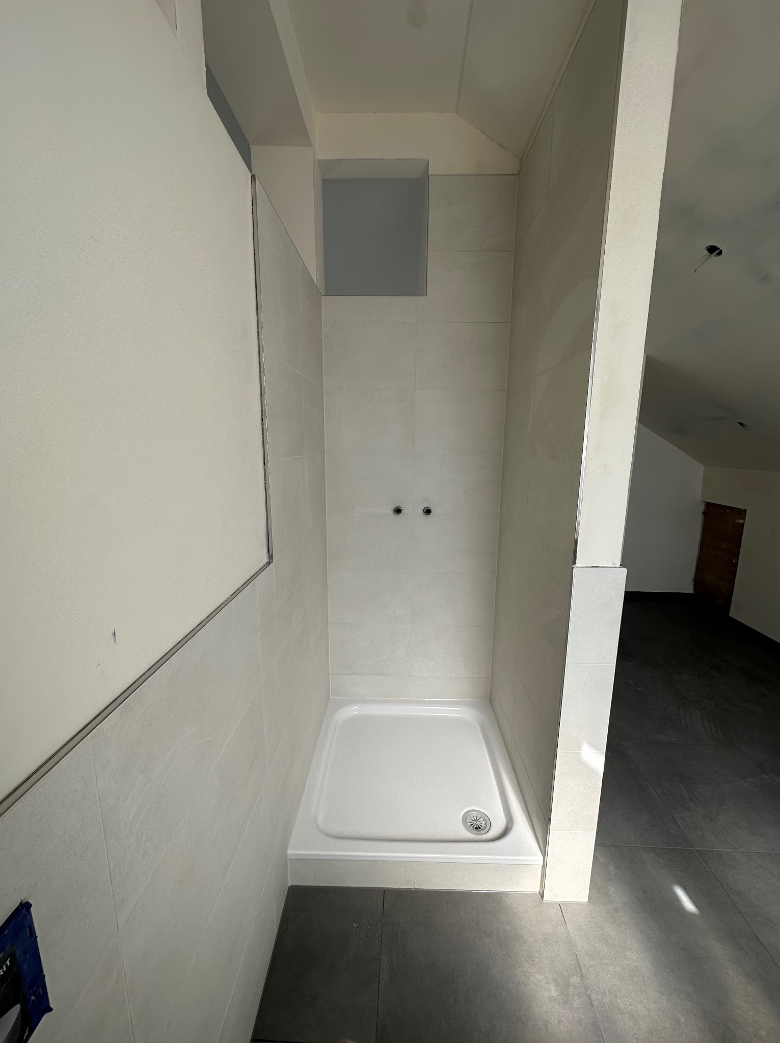 Wnętrze kabiny prysznicowej z białym brodzikiem i jasnymi płytkami, widoczne dwa wystające przyłącza wodne, surowy stan wykończenia, ciemna podłoga z płytek.