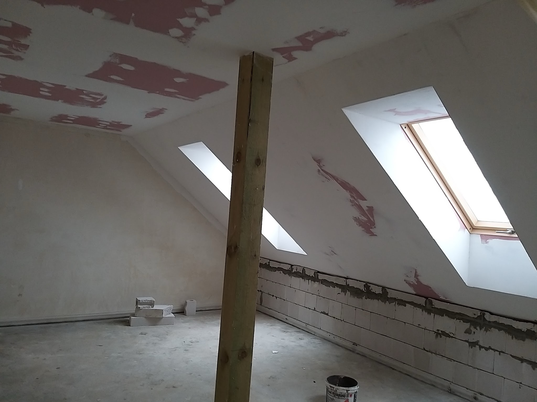 Poddasze w trakcie remontu: ściany z bloków połączonych zaprawą, częściowo pokryte szpachlą, sufit z widocznymi łatami szpachli, drewniany słup podtrzymujący, okna dachowe, na podłodze resztki...