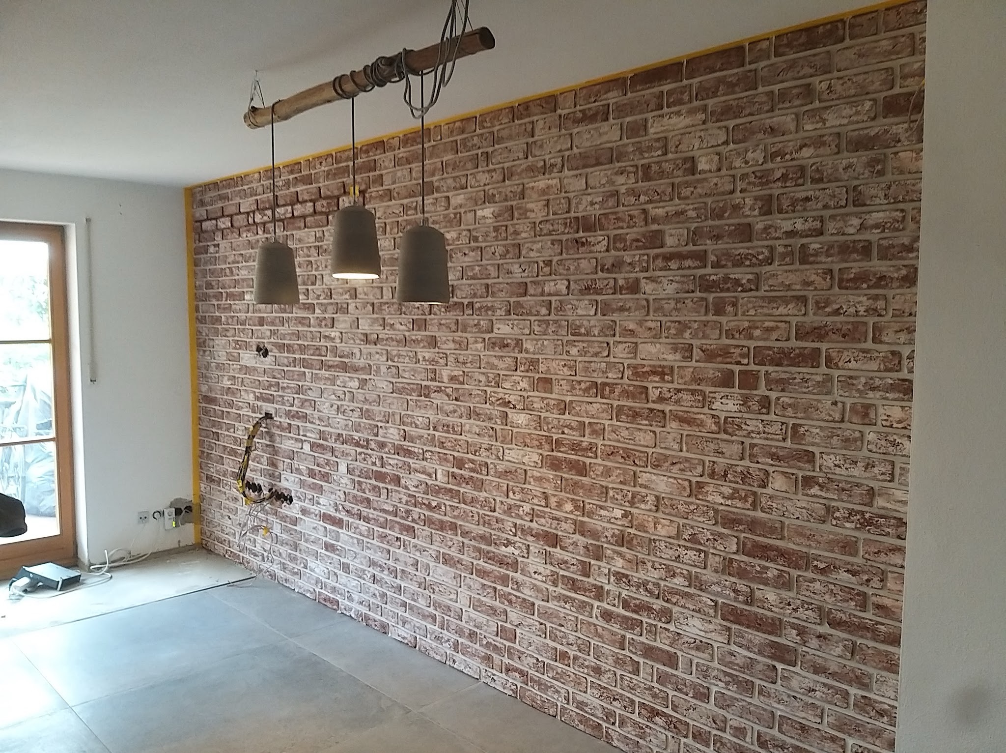 Ściana wykończona cegłą dekoracyjną w odcieniach czerwieni i bieli, z widocznymi przewodami elektrycznymi wystającymi ze ściany i trzema szarymi lampami wiszącymi na drewnianej belce, żółta taśma...