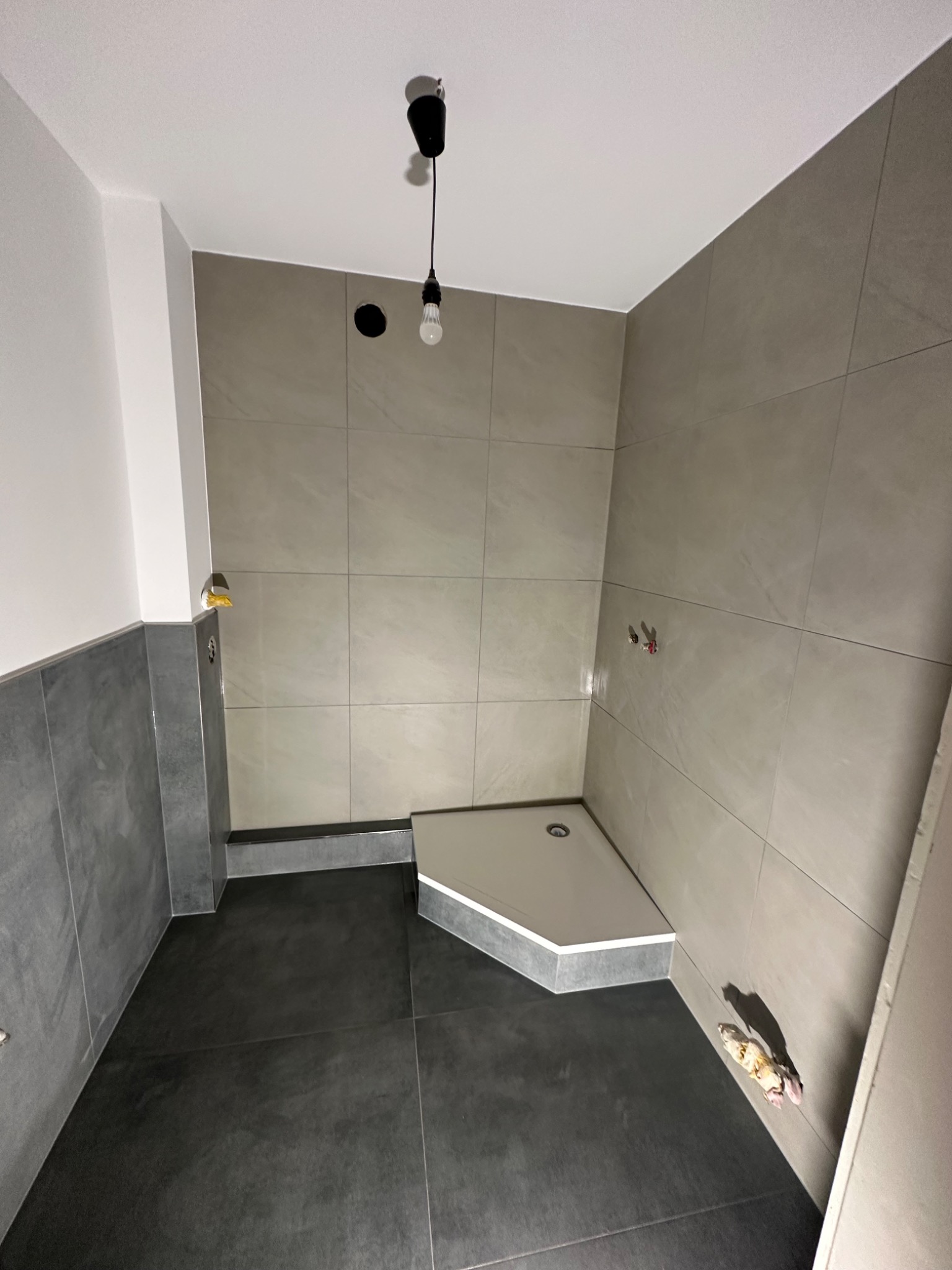 Nowoczesna łazienka w trakcie wykańczania: szare płytki na ścianach, czarna podłoga, biały brodzik. Widoczne niedokończone elementy instalacji i surowa żarówka.