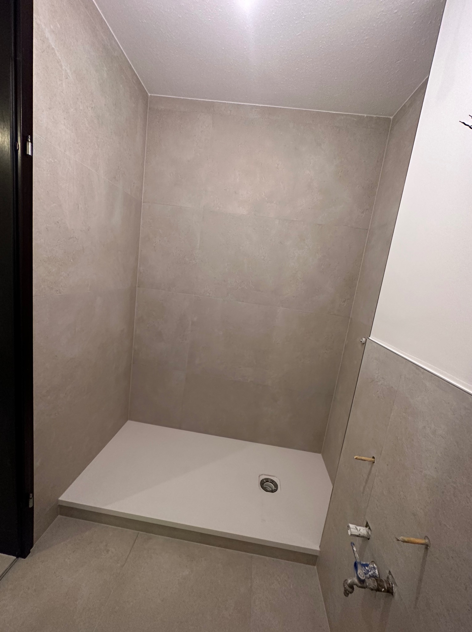 Wykończona kabina prysznicowa z beżowymi płytkami, widoczne niedokończone przyłącza wodne. Minimalistyczny design, jasne oświetlenie.