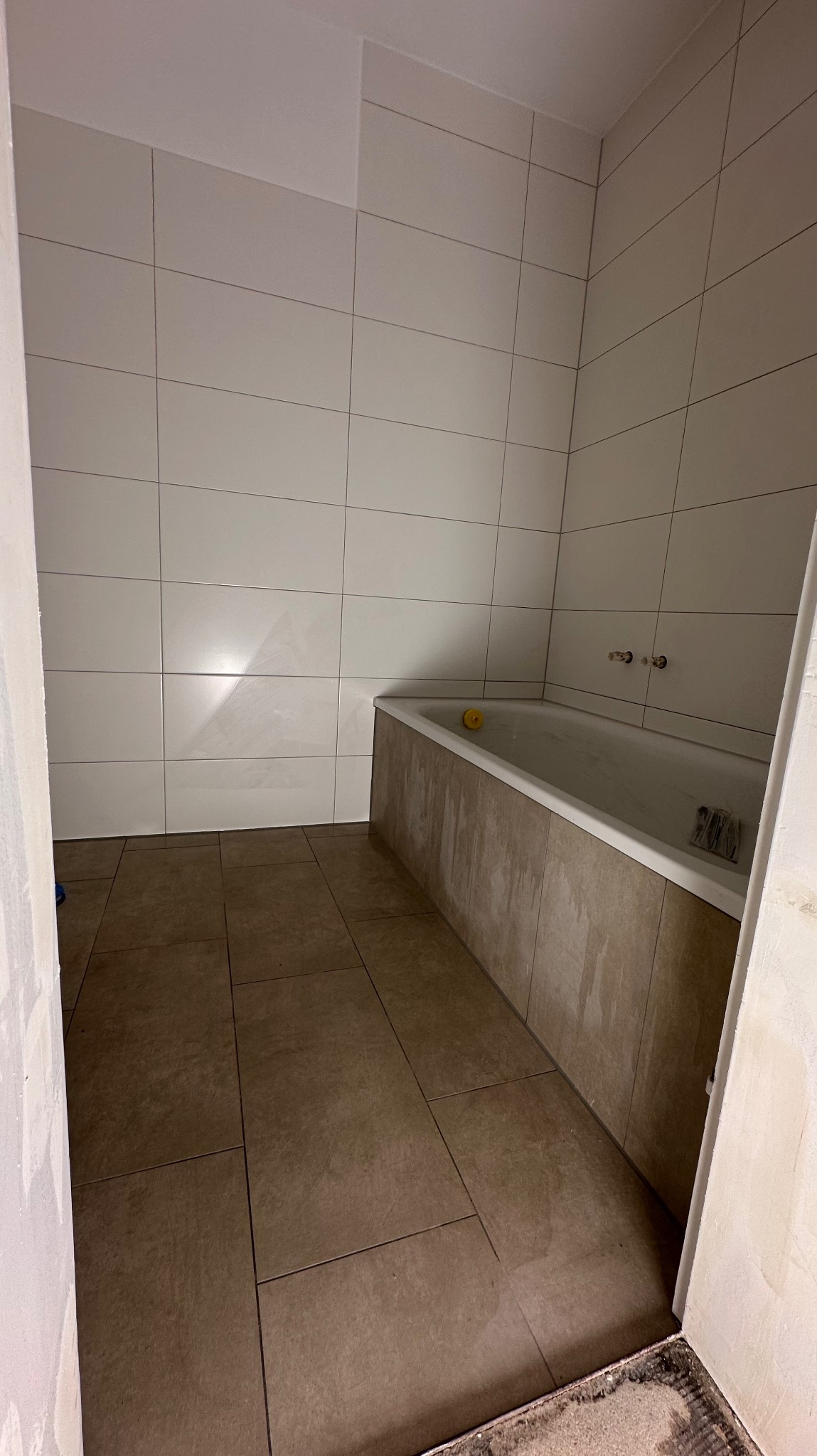 Łazienka w trakcie wykończenia: ułożone brązowe płytki podłogowe i białe płytki ścienne, obudowana wanna. Widoczne niedokończone wykończenia.