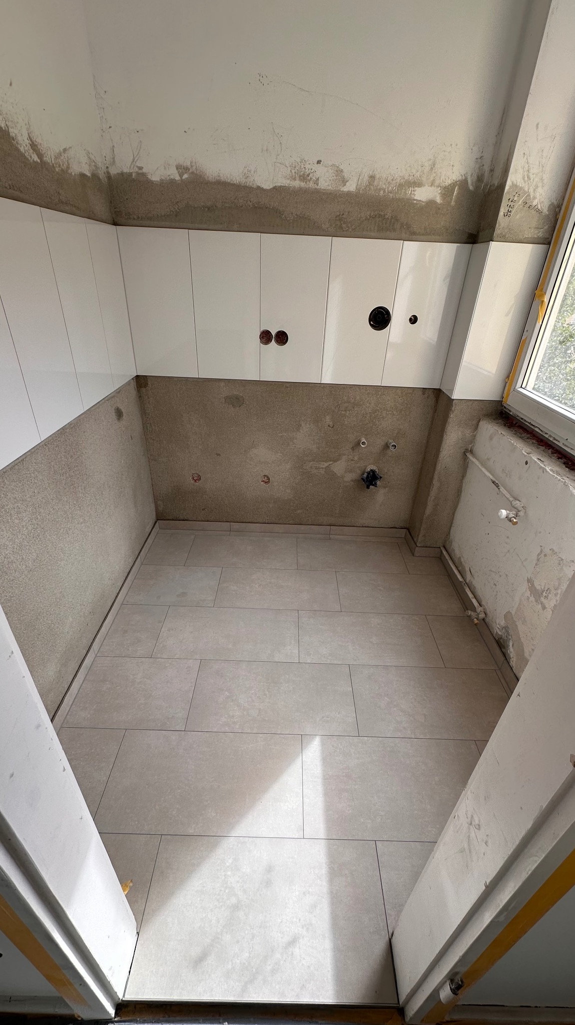 Łazienka w trakcie prac: ułożona podłoga z płytek, częściowo położone białe płytki na ścianach, widoczne instalacje elektryczne i hydrauliczne. Okno z prawej strony.