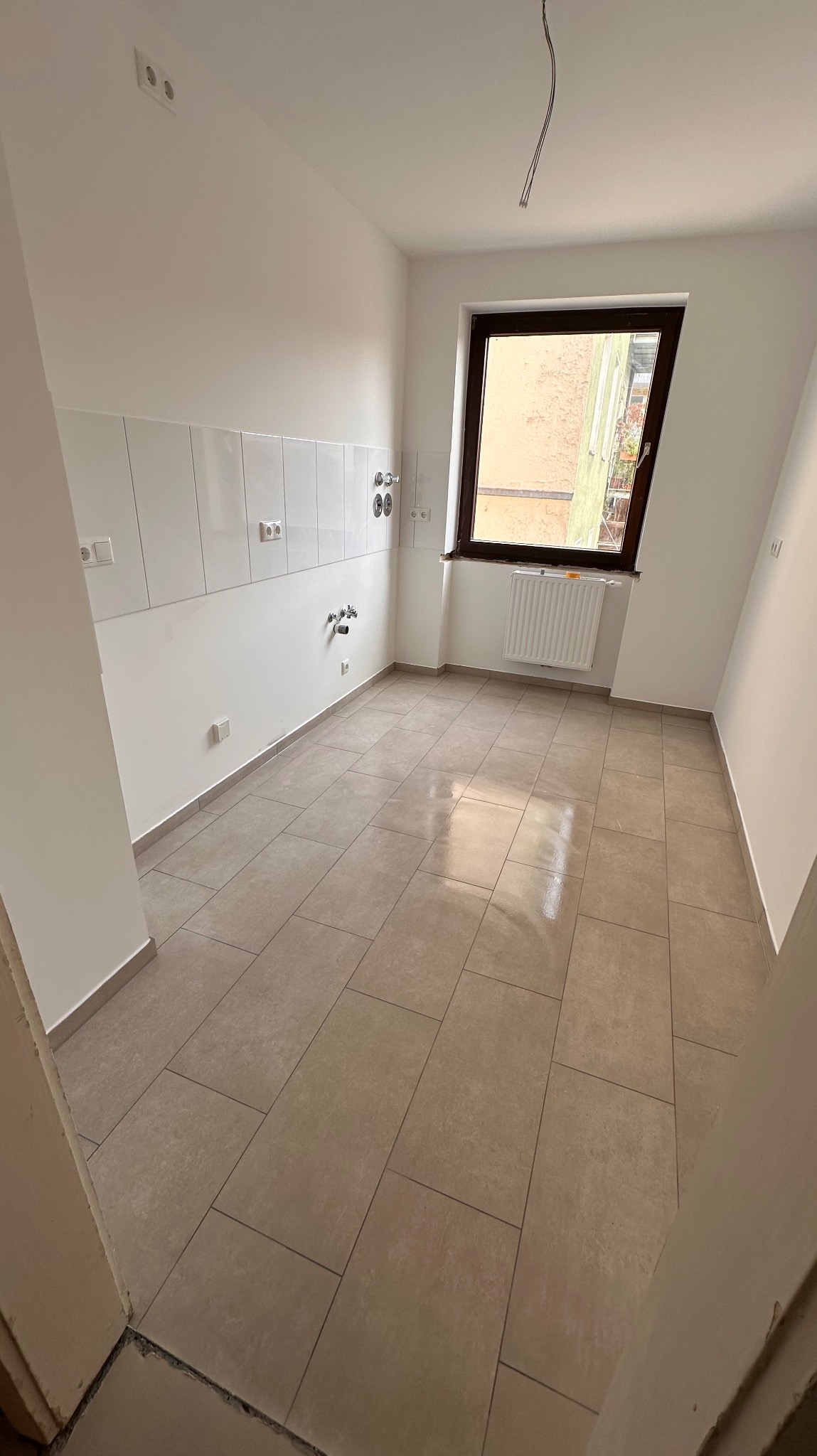 Jasna łazienka po remoncie: beżowe płytki podłogowe, białe ściany z fragmentem glazury, brązowe okno. Widoczne przyłącza wodne i elektryczne.