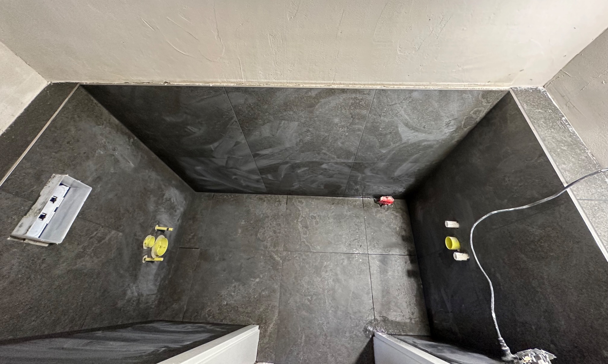 Ciemne, matowe płytki w kabinie prysznicowej w trakcie montażu. Widoczne żółte elementy instalacji wodnej i narzędzia glazurnicze. Górny kadr, surowy stan wykończenia.