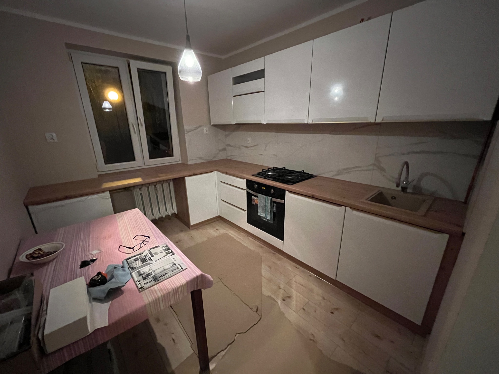 Nowoczesna, biała zabudowa kuchenna z drewnianym blatem, widok z boku, na stole leżą okulary i katalog meblowy IKEA.