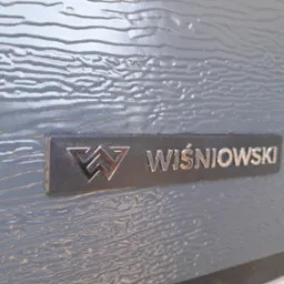 Szczegółowe zbliżenie na panel bramy garażowej marki Wiśniowski we Wrocławiu, prezentujący fakturę imitującą drewno i metalowe logo producenta.