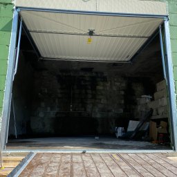 DM SERWIS - Montaż segmentowej bramy garażowej: widok od wewnątrz na częściowo otwartą, białą bramę z widoczną konstrukcją montażową na tle surowego wnętrza garażu z zielonymi ścianami zewnętrznymi.