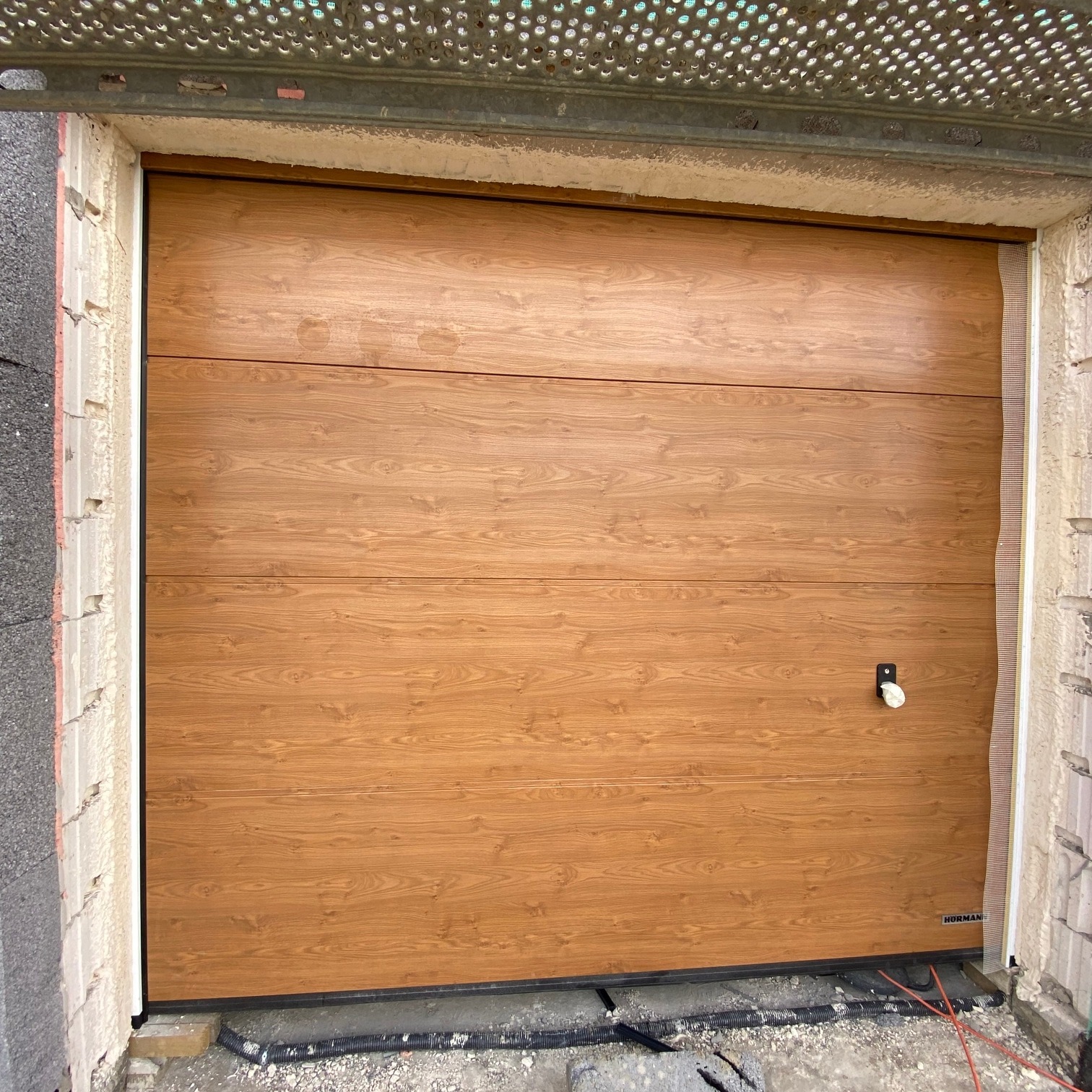 Zamknięta segmentowa brama garażowa w kolorze jasnego dębu, z widocznym zamkiem i logo producenta, zamontowana w surowym otworze garażowym z widocznymi elementami konstrukcyjnymi.