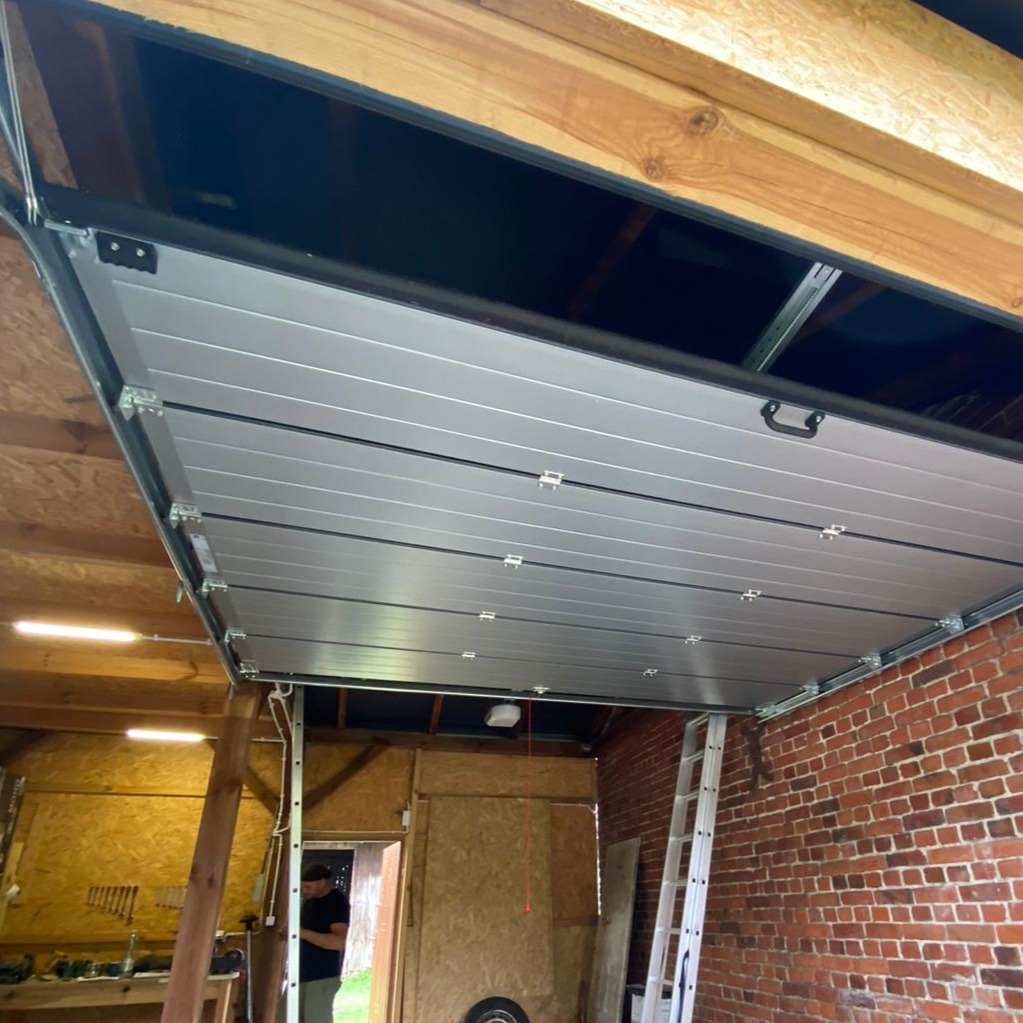 Montaż segmentowej bramy garażowej z widocznymi prowadnicami i elementami mocującymi do stropu, ceglana ściana garażu w tle.