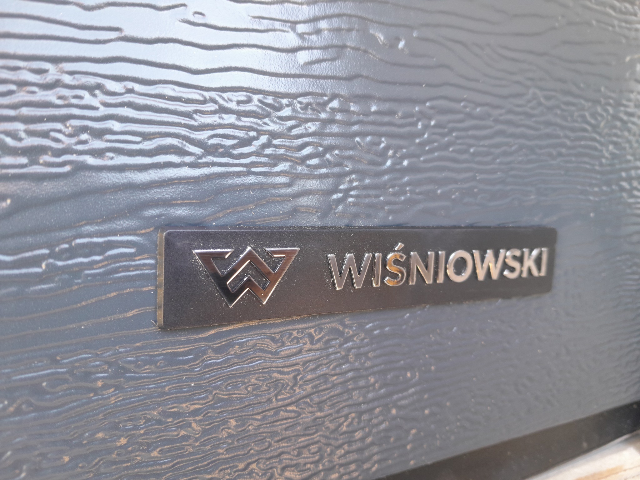 Szczegółowe zbliżenie na panel bramy garażowej marki Wiśniowski we Wrocławiu, prezentujący fakturę imitującą drewno i metalowe logo producenta.