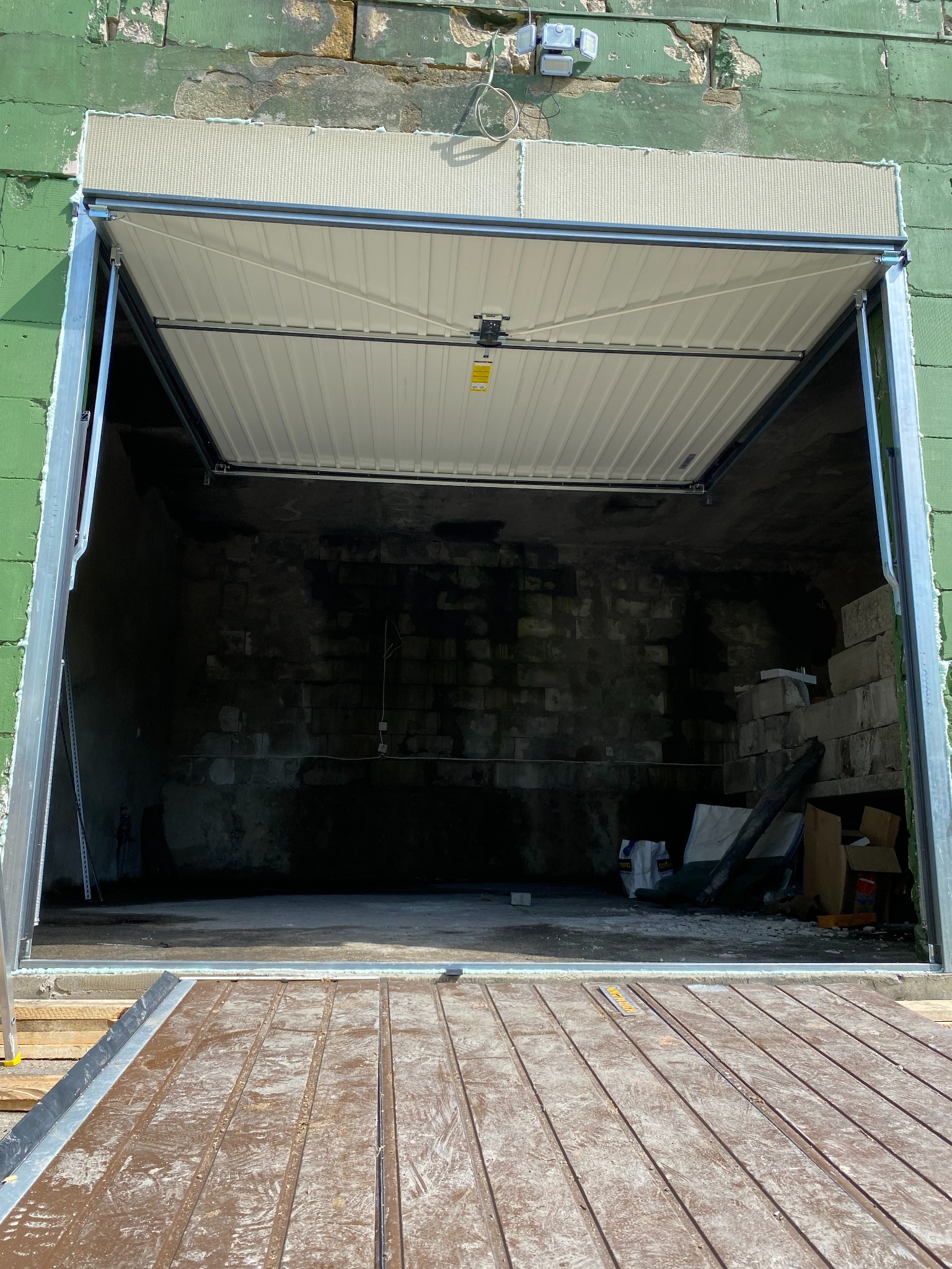 Montaż segmentowej bramy garażowej: widok od wewnątrz na częściowo otwartą, białą bramę z widoczną konstrukcją montażową na tle surowego wnętrza garażu z zielonymi ścianami zewnętrznymi.