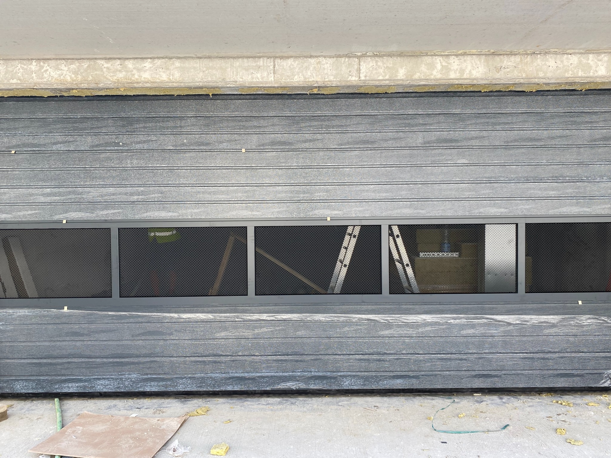 Częściowo otwarta, szara brama segmentowa z rzędem okienek z siatką, widoczne elementy montażowe i drabina wewnątrz garażu.