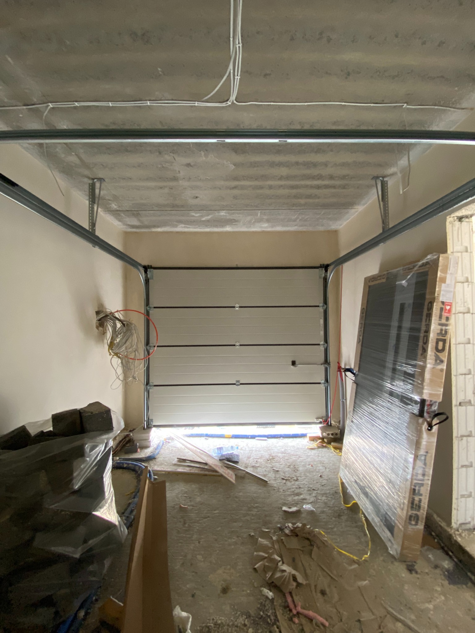 Wnętrze garażu w trakcie montażu segmentowej bramy, widoczne przewody elektryczne wystające ze ściany, zapakowana brama oparta o ścianę, surowy betonowy sufit.