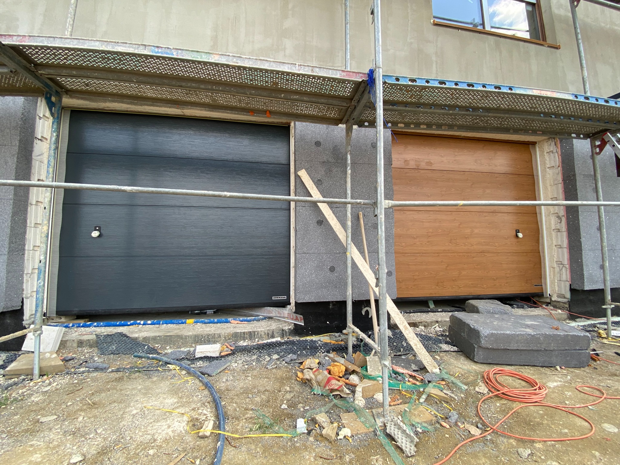 Dwie segmentowe bramy garażowe: szara i drewnopodobna, w trakcie montażu, widoczne rusztowanie i materiały budowlane na placu budowy.