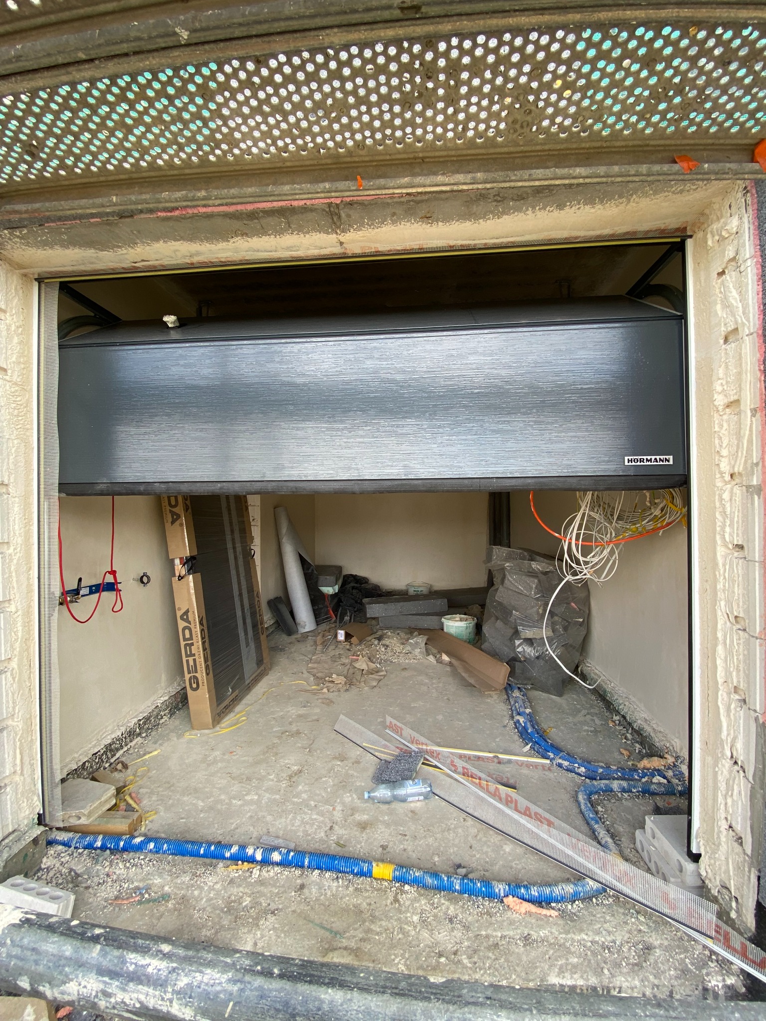Częściowo opuszczona, grafitowa brama garażowa marki Hormann, widoczna z wnętrza garażu w trakcie prac wykończeniowych, z widocznymi przewodami elektrycznymi i materiałami budowlanymi.