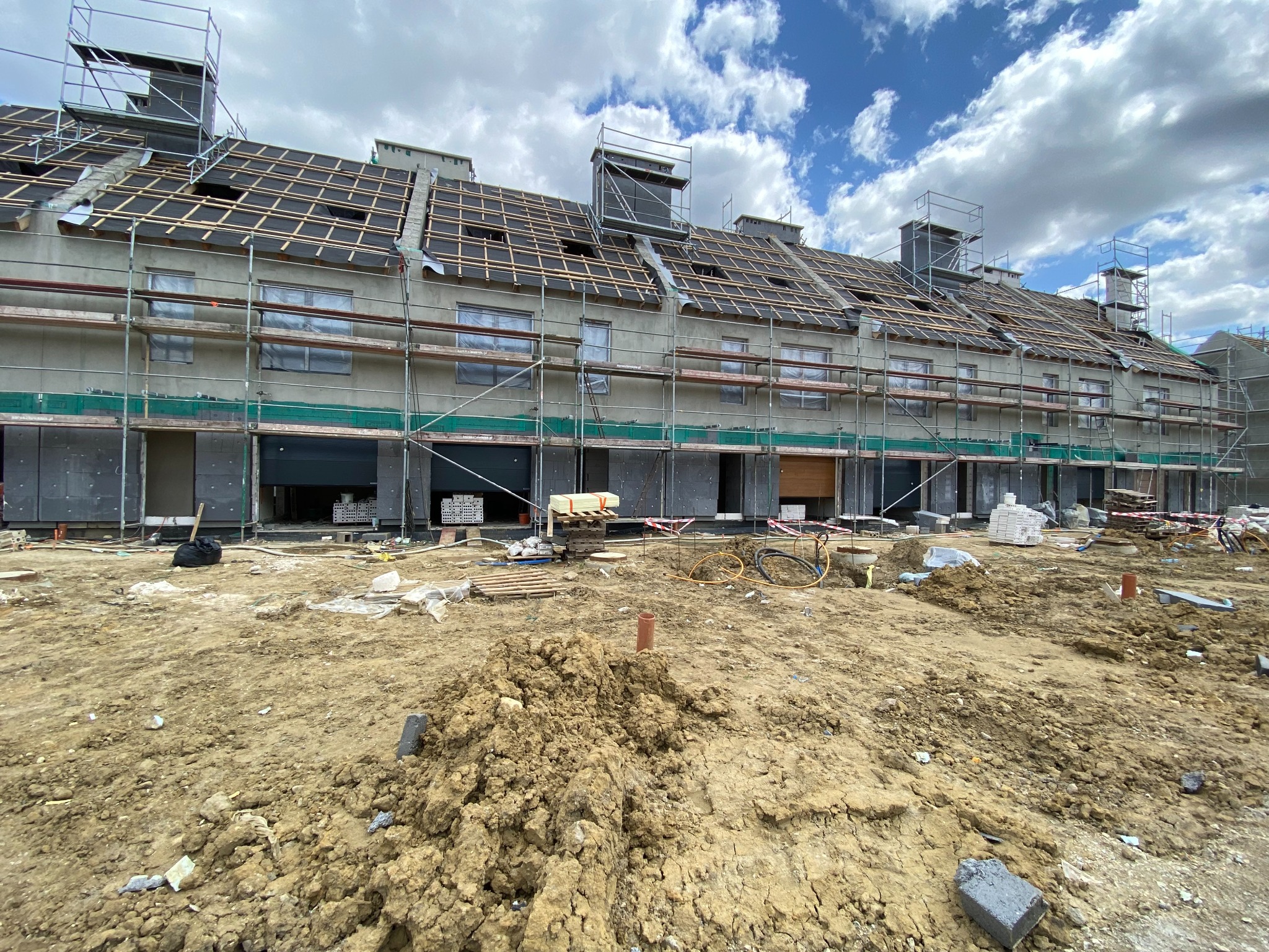 Wielorodzinny budynek w trakcie budowy, widoczne rusztowania, niezadaszony dach z drewnianą konstrukcją, teren budowy z hałdą ziemi na pierwszym planie i rowerem opartym o ścianę budynku.