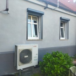 Pw Reda - Szara ściana budynku z dwoma oknami z szarymi roletami zewnętrznymi, klimatyzator Aiwa na metalowym stelażu oraz krzew hortensji.