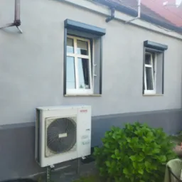 Szara ściana budynku z dwoma oknami z szarymi roletami zewnętrznymi, klimatyzator Aiwa na metalowym stelażu oraz krzew hortensji.