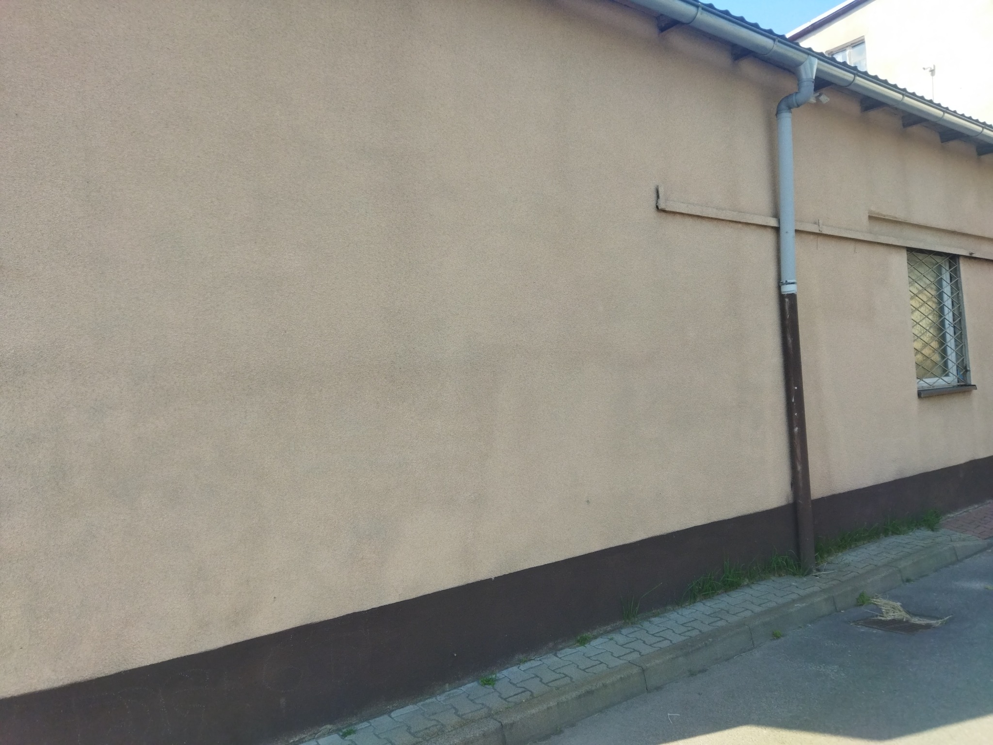 Jasnożółta elewacja budynku z widocznym fragmentem orynnowania, pomalowanym na brązowo cokołem i fragmentem chodnika z kostki brukowej.
