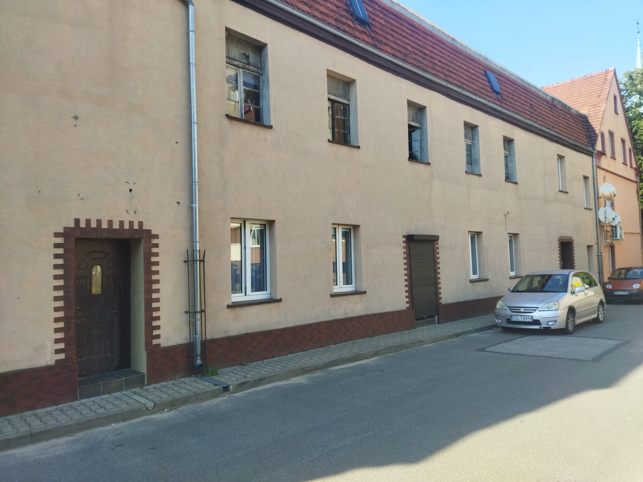 Długi budynek mieszkalny z beżową elewacją i ciemnoczerwoną ceglaną podstawą, widoczny z poziomu ulicy, z zaparkowanym samochodem osobowym przed budynkiem.